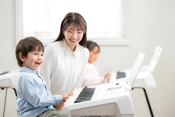 A music lesson, with a student  playing the keyboard while a teacher watches over them while sitting next to them